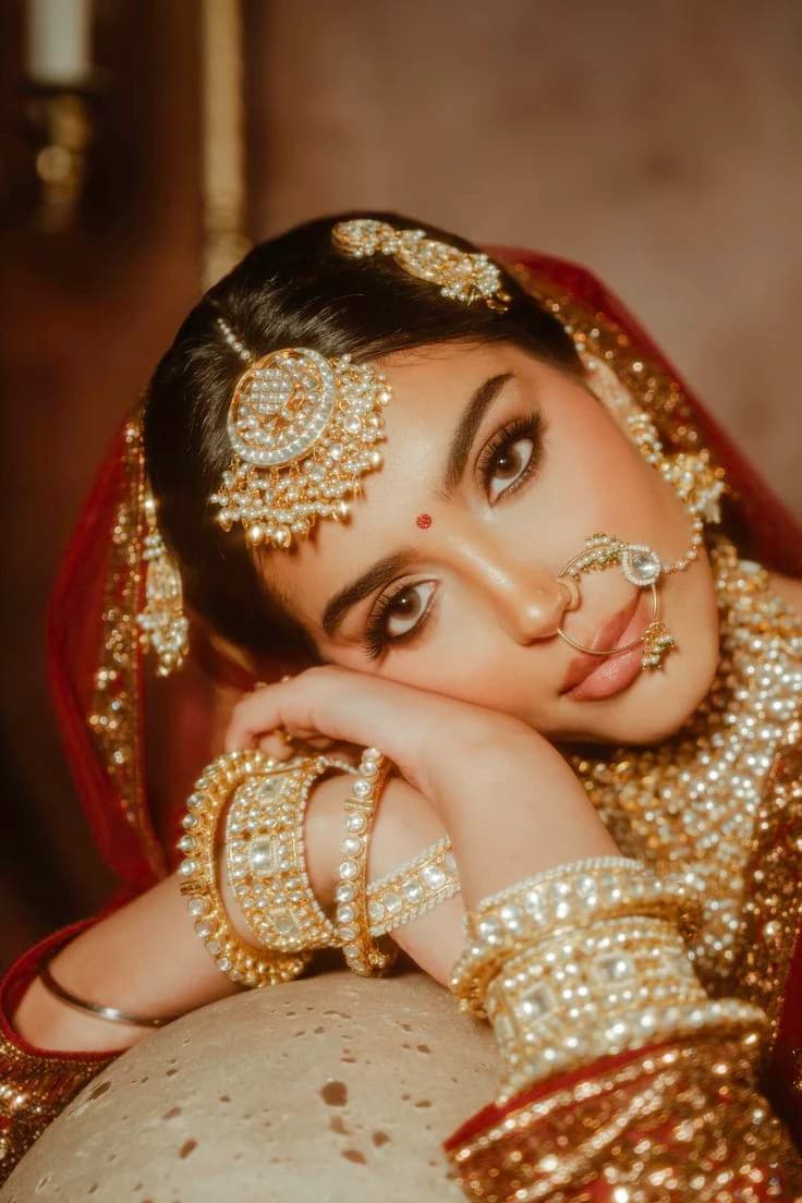 Bride resting her cheek on her hand, wearing a pearl maang tikka, nose ring and stacked gold bangles