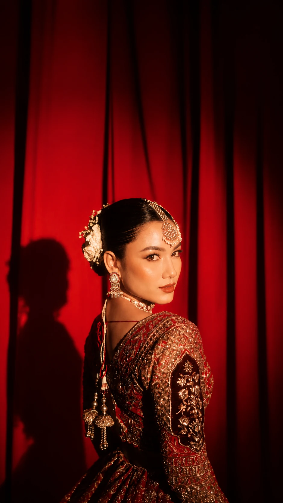 Bride in a maroon lehenga with a gold maang tikka and pearl earrings, looking over her shoulder